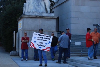 When I first made the scene and took this picture, Justice (with sign) didn’t realize (and neither did I, frankly) he was standing beneath a statue depicting the “Spirit of Justice,” as the engraving above his head here reads. On the other side, flanking the steps on the right up to the entrance to the Rayburn building, is the “Majesty of Law” — the marble statues were sculpted by C. Paul Jennewein .