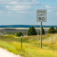 Curved highway with 80 mph speed limit sign amid grassy landscape and distant trees