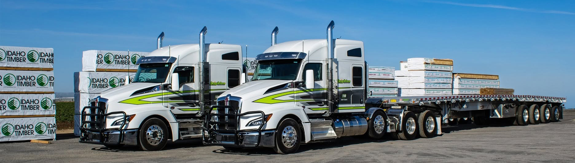 Two white and lime green semi-trucks with trailers loaded with stacked materials at a logistics facility