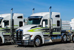 Two white and lime green semi-trucks with trailers loaded with stacked materials at a logistics facility