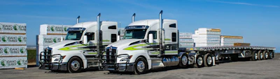 Two white and lime green semi-trucks with trailers loaded with stacked materials at a logistics facility