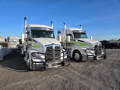 Two white commercial semi-trucks with blue and green striping parked in a fleet yard