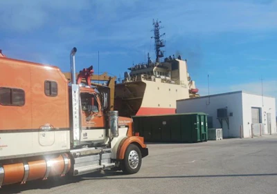 Orange and white truck parked at industrial port facility with cargo ship docked in background