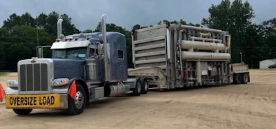 Heavy-duty Peterbilt tractor-trailer truck hauling oversized industrial equipment on dirt lot with oversize load warning sign
