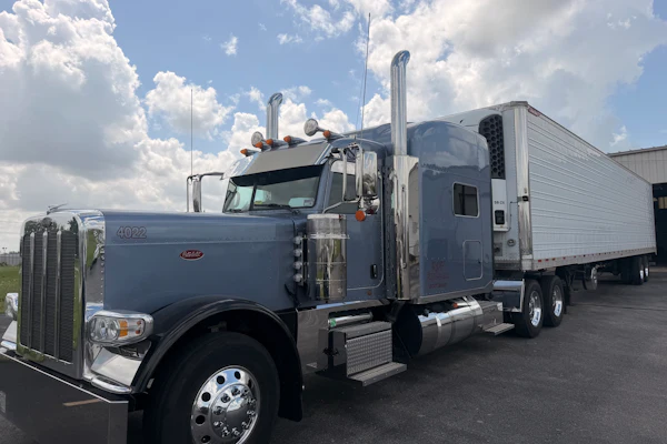 Gray Peterbilt semi-truck with white refrigerated trailer parked in lot under cloudy sky
