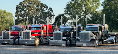 Fleet of red, black, and gray semi-trucks lined up in a row on a parking lot with trees in the background