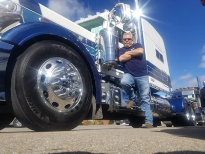 Professional truck driver posing confidently next to his blue and white semi-truck on a sunny day