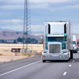 Turquoise semi-truck leading traffic on highway with mountains and transmission tower in background