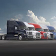 Row of five commercial semi-trucks in navy, white, red, and blue colors parked on asphalt against a cloudy sky