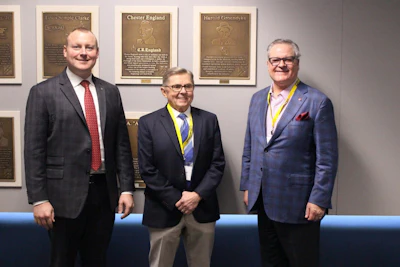Three men in business attire stand together at a formal event in front of bronze commemorative plaques on a wall.