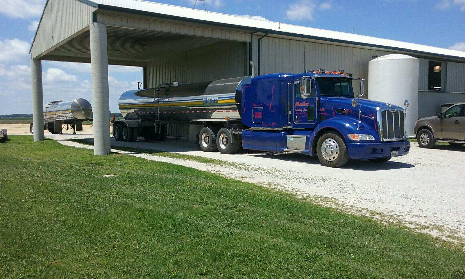 Blue Peterbilt tanker truck parked under metal canopy with white storage tank nearby