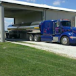 Blue Peterbilt tanker truck parked under metal canopy with white storage tank nearby