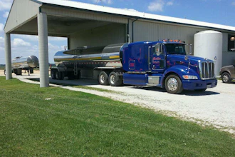 Blue Peterbilt tanker truck parked under metal canopy with white storage tank nearby