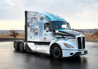 White and light blue Peterbilt semi-truck with nature-themed cab wrap parked on asphalt with rural landscape background