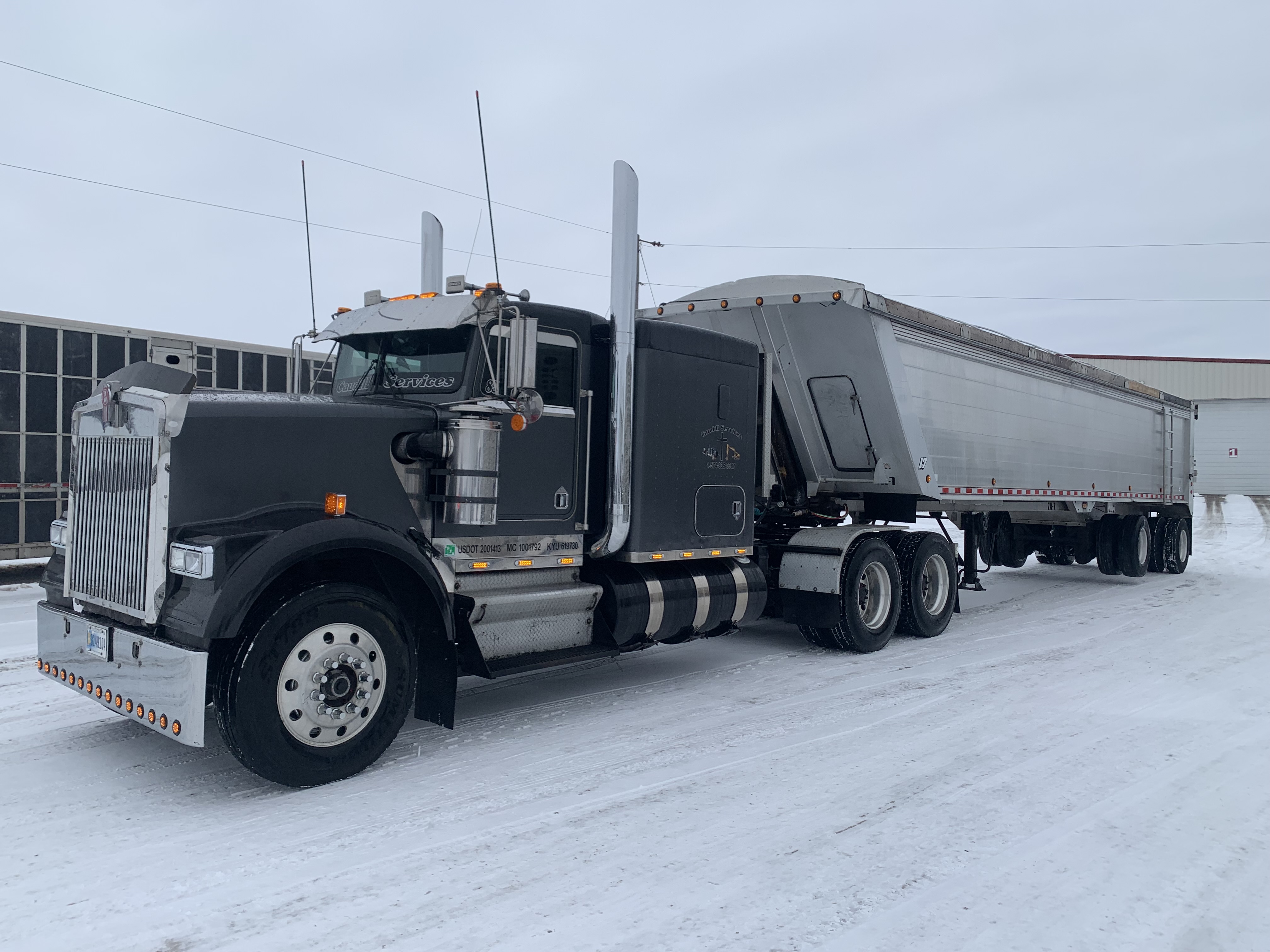 Black Peterbilt semi-truck with silver grain trailer parked in snowy industrial lot