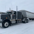 Black Peterbilt semi-truck with silver grain trailer parked in snowy industrial lot