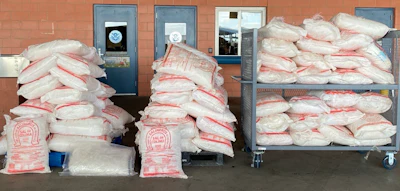 Large quantities of packaged flour or bulk commodity stacked in warehouse with industrial equipment visible in background