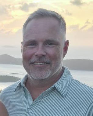 Professional headshot of a middle-aged man smiling at the camera with a coastal landscape background. Headshot