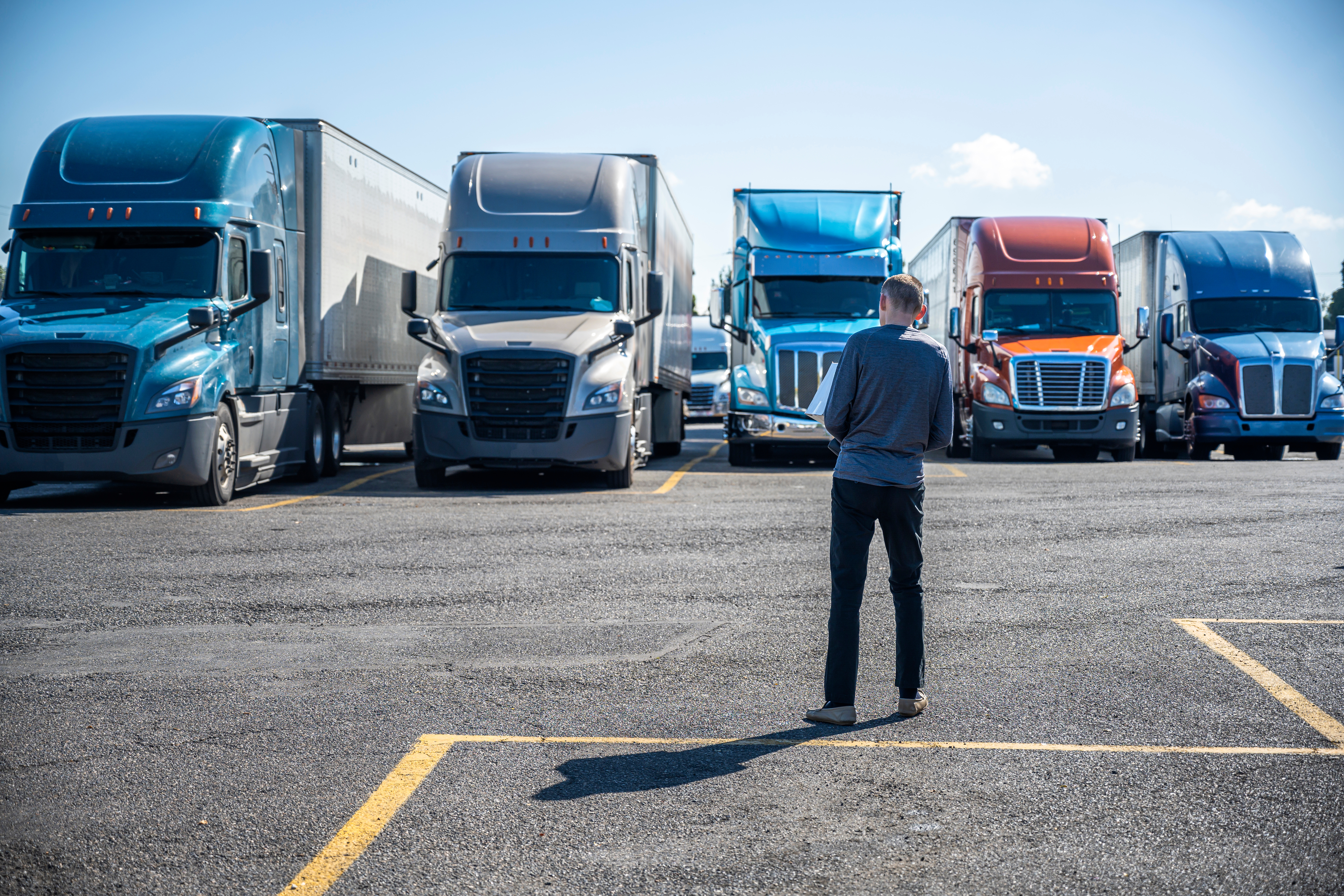 Businessman standing in parking lot facing row of commercial semi-trucks in various colors under clear sky