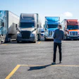 Businessman standing in parking lot facing row of commercial semi-trucks in various colors under clear sky
