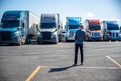 Businessman standing in parking lot facing row of commercial semi-trucks in various colors under clear sky