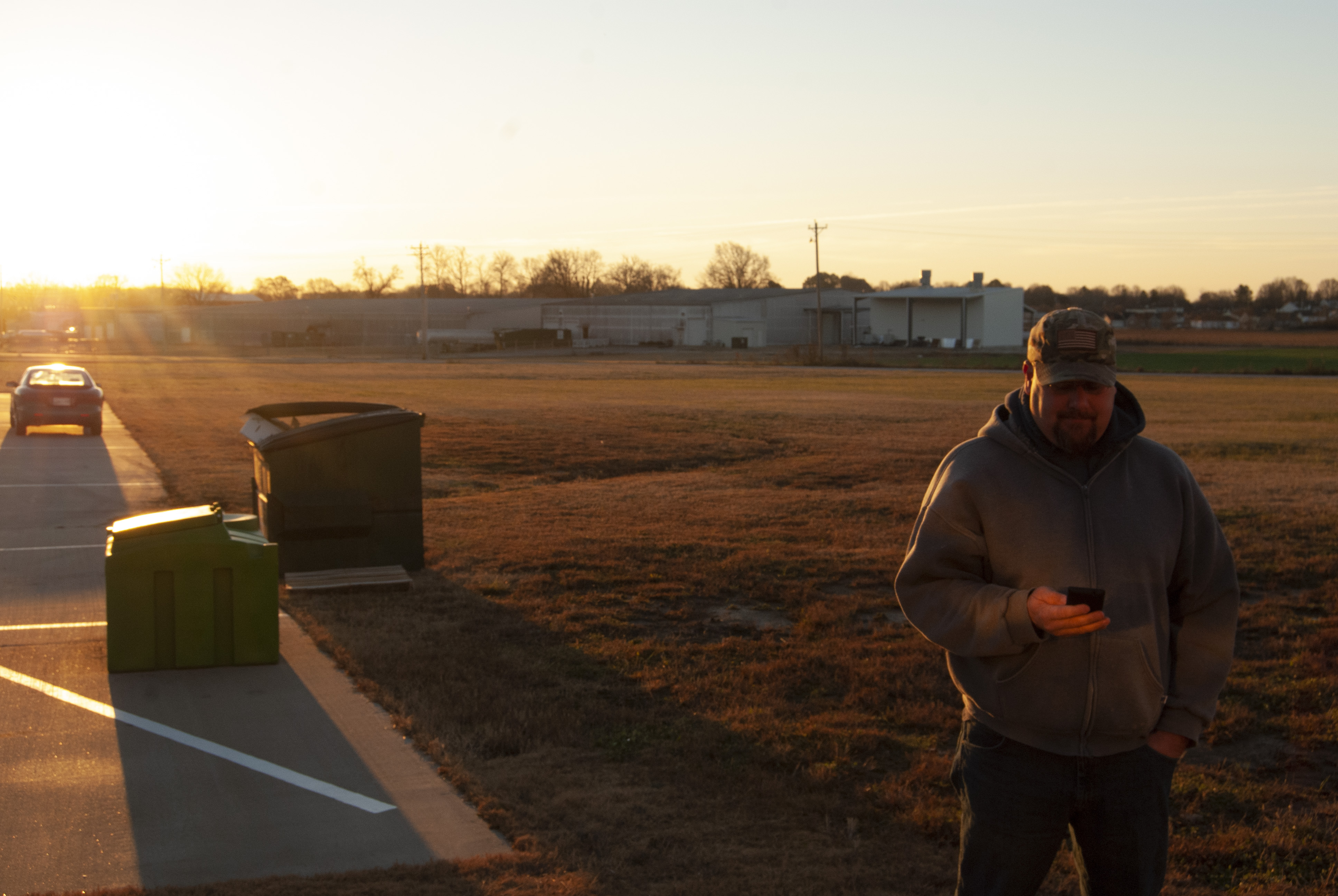 Person standing in rural area at sunset with vehicles and farm buildings in background