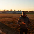 Person standing in rural area at sunset with vehicles and farm buildings in background