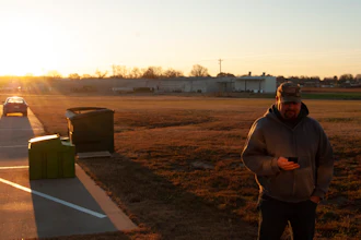 Person standing in rural area at sunset with vehicles and farm buildings in background