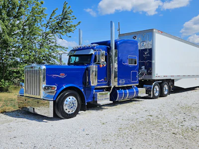 Bright blue Peterbilt semi-truck with white refrigerated trailer parked on gravel against blue sky