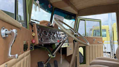 Vintage truck interior with tan dashboard, steering wheel, and circular gauges overlooking other classic vehicles outside