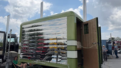 Green and brown auto transport trailer loaded with stacked vehicles at an outdoor facility under cloudy sky