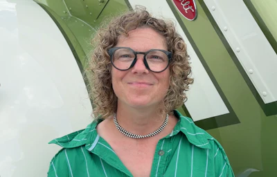Person with curly hair and glasses wearing a green striped shirt and beaded necklace