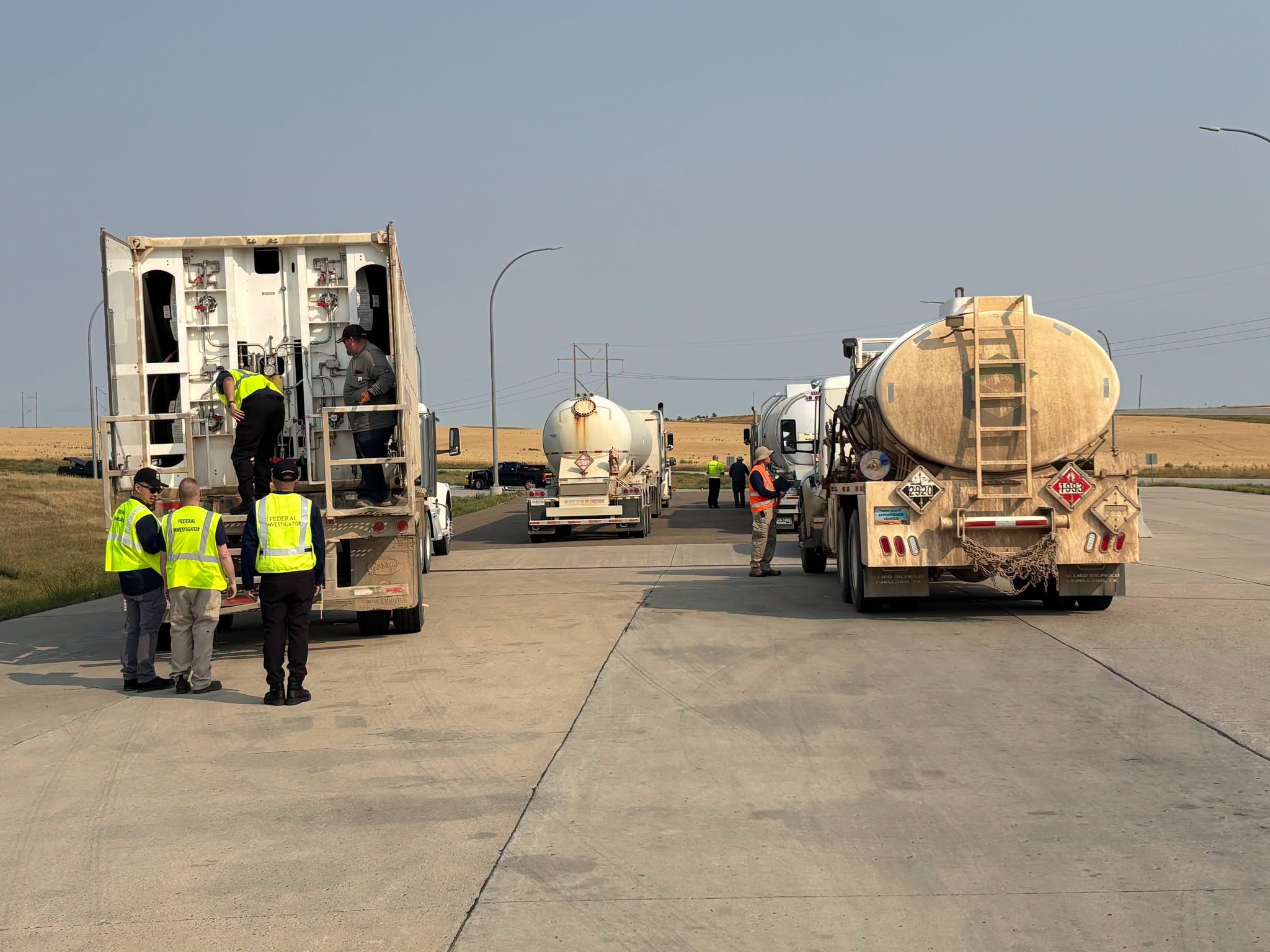 Workers in safety vests inspect tanker trucks on a highway during daytime operations.