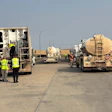 Workers in safety vests inspect tanker trucks on a highway during daytime operations.