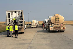 Workers in safety vests inspect tanker trucks on a highway during daytime operations.