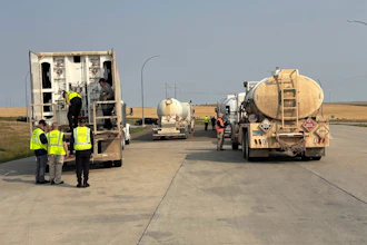 Workers in safety vests inspect tanker trucks on a highway during daytime operations.