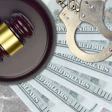 Gavel, handcuffs, and hundred-dollar bills arranged on concrete surface representing law, justice, and financial crime.