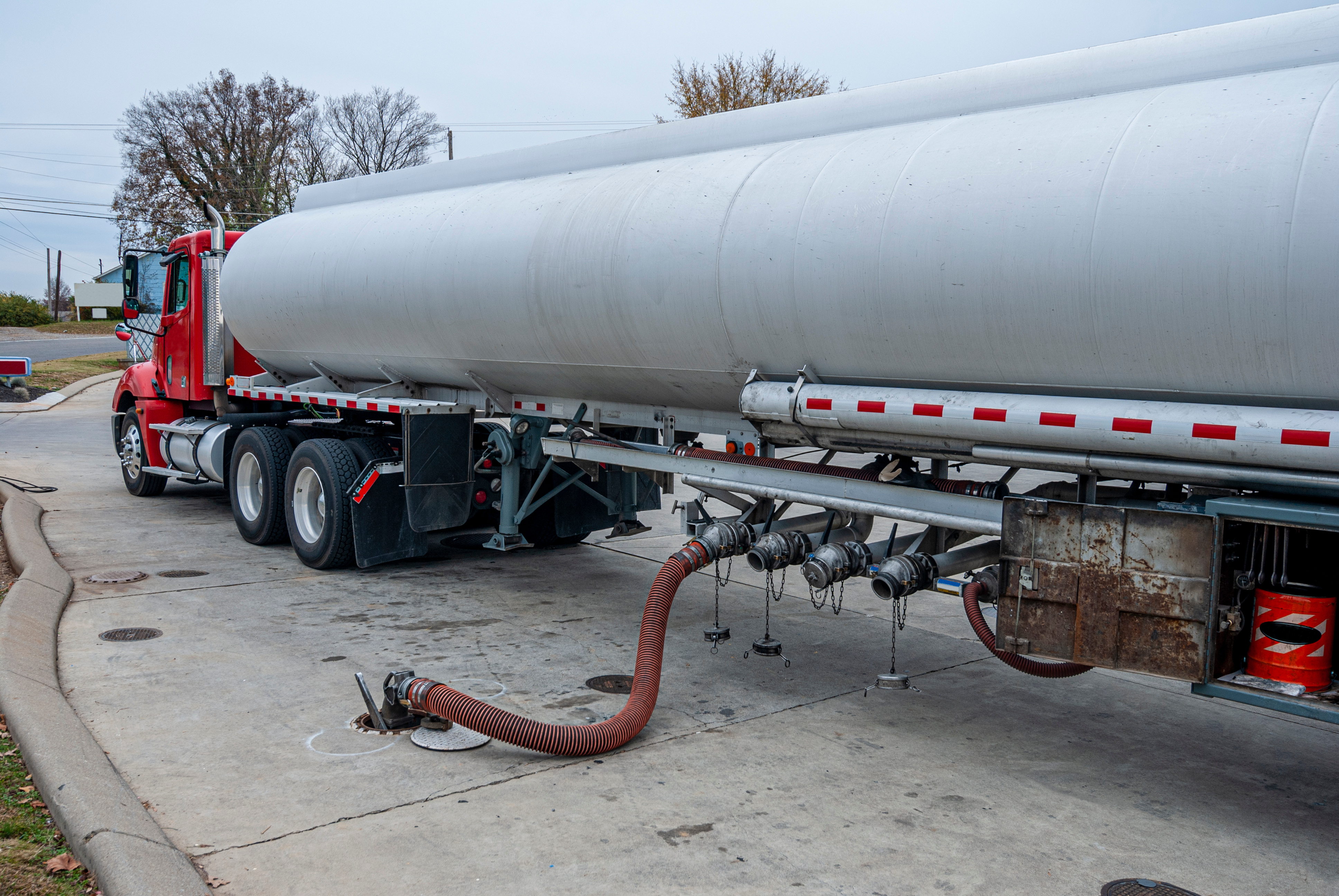 Red tanker truck with white cylindrical tank parked at industrial facility with discharge hose