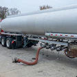 Red tanker truck with white cylindrical tank parked at industrial facility with discharge hose