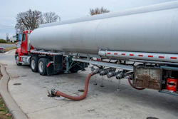 Red tanker truck with white cylindrical tank parked at industrial facility with discharge hose