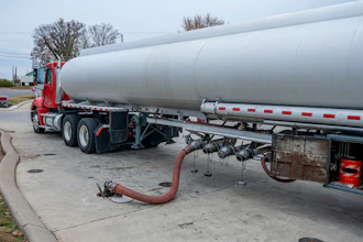 Red tanker truck with white cylindrical tank parked at industrial facility with discharge hose