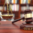 Wooden gavel with 'GUILTY' engraved on base beside brass scales of justice in law library setting