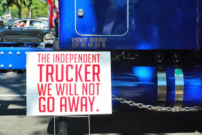 Blue commercial truck displaying white protest sign with red text reading 'The Independent Trucker We Will Not Go Away'