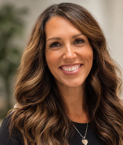 Professional headshot of smiling woman with long brown wavy hair wearing black top and heart pendant necklace