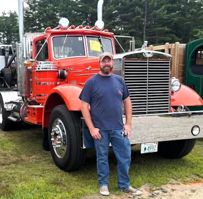 Man in navy shirt and cap standing in front of restored red vintage semi-truck at outdoor truck show