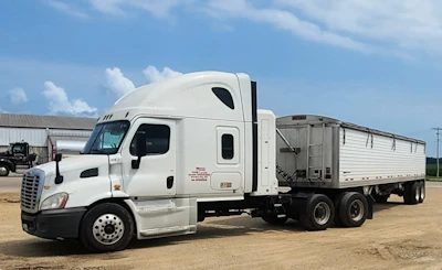 White Freightliner semi-truck with grain trailer parked on dirt lot under blue sky