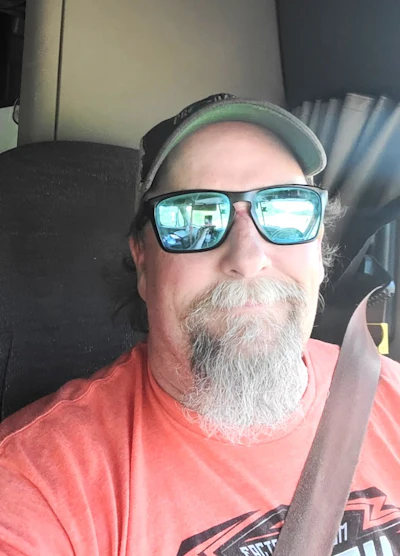 Middle-aged man with gray beard wearing green cap, blue-tinted sunglasses, and coral shirt inside a vehicle
