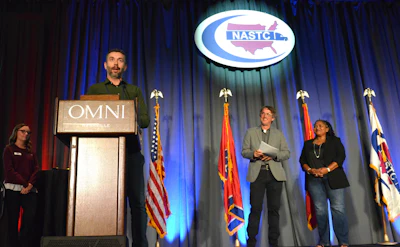 Speaker at OMNI podium during NASCO conference with attendees and flags on stage