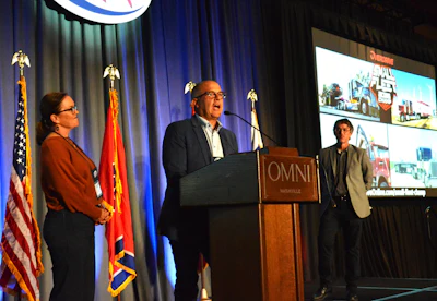Professional speaking at OMNI Nashville conference podium with two attendees and presentation screens displaying equipment imagery