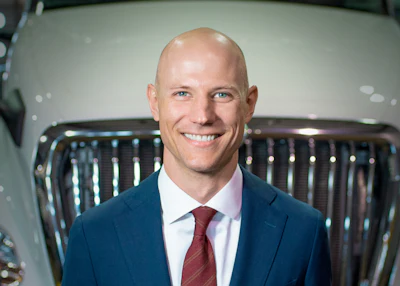 Professional man in blue blazer and burgundy tie smiling at camera with vehicle grille background